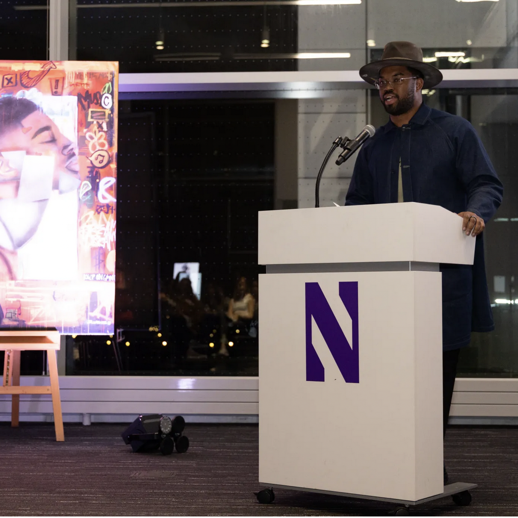 Dwight White stands at white podium detailed with purple N next to painting of man's face in side profile