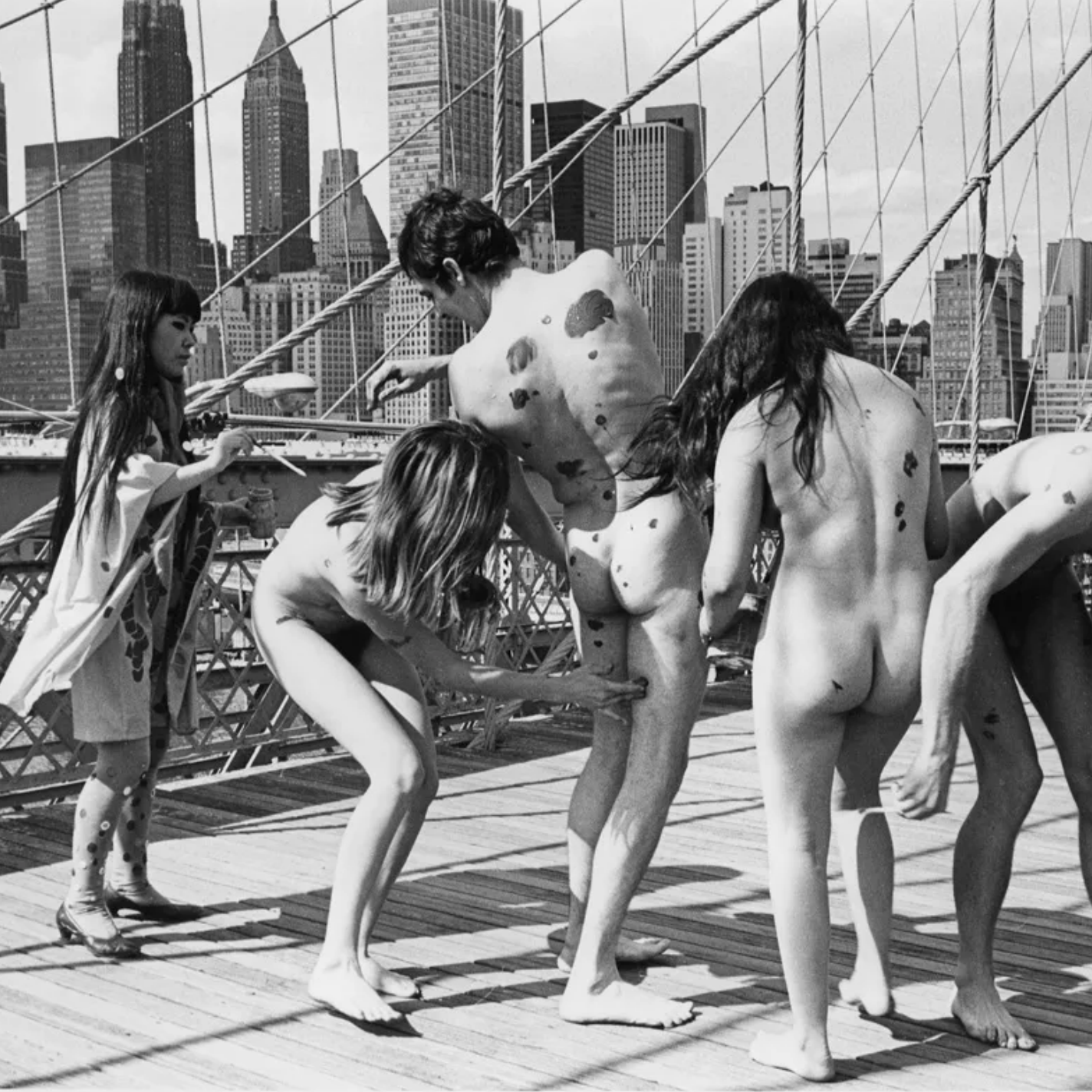several nude figures fingerpaint one another on bridge with city skyline in background