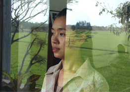A woman looks out from behind a window, which reflects a green field. 