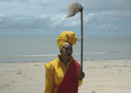 closeup of a Black person with dark skin tone, in yellow and red garment and head wrap, holding a staff and on the beach