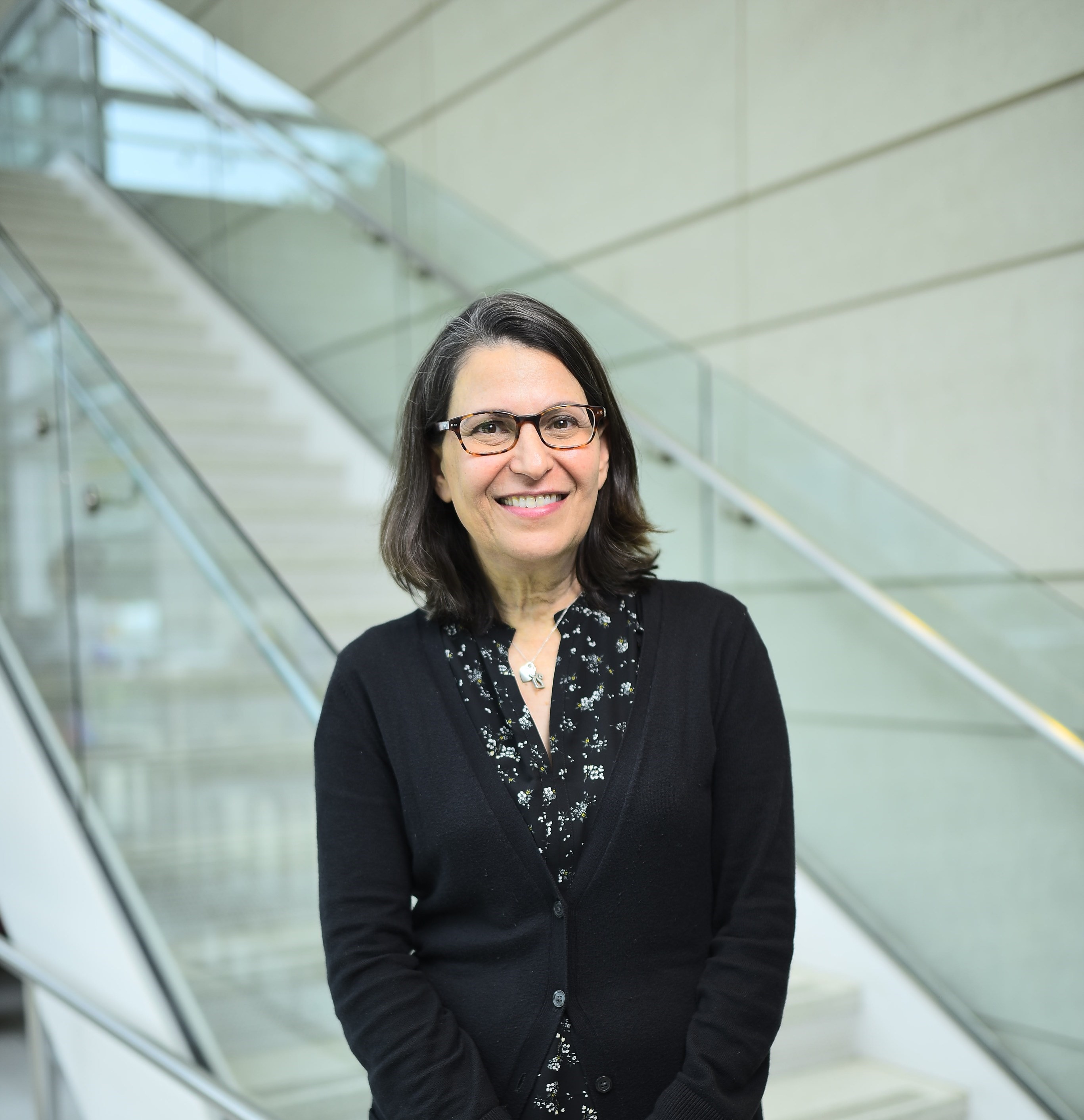 photographic portrait of white woman with glasses and shoulder length brown hair, standing in front of a staircase 