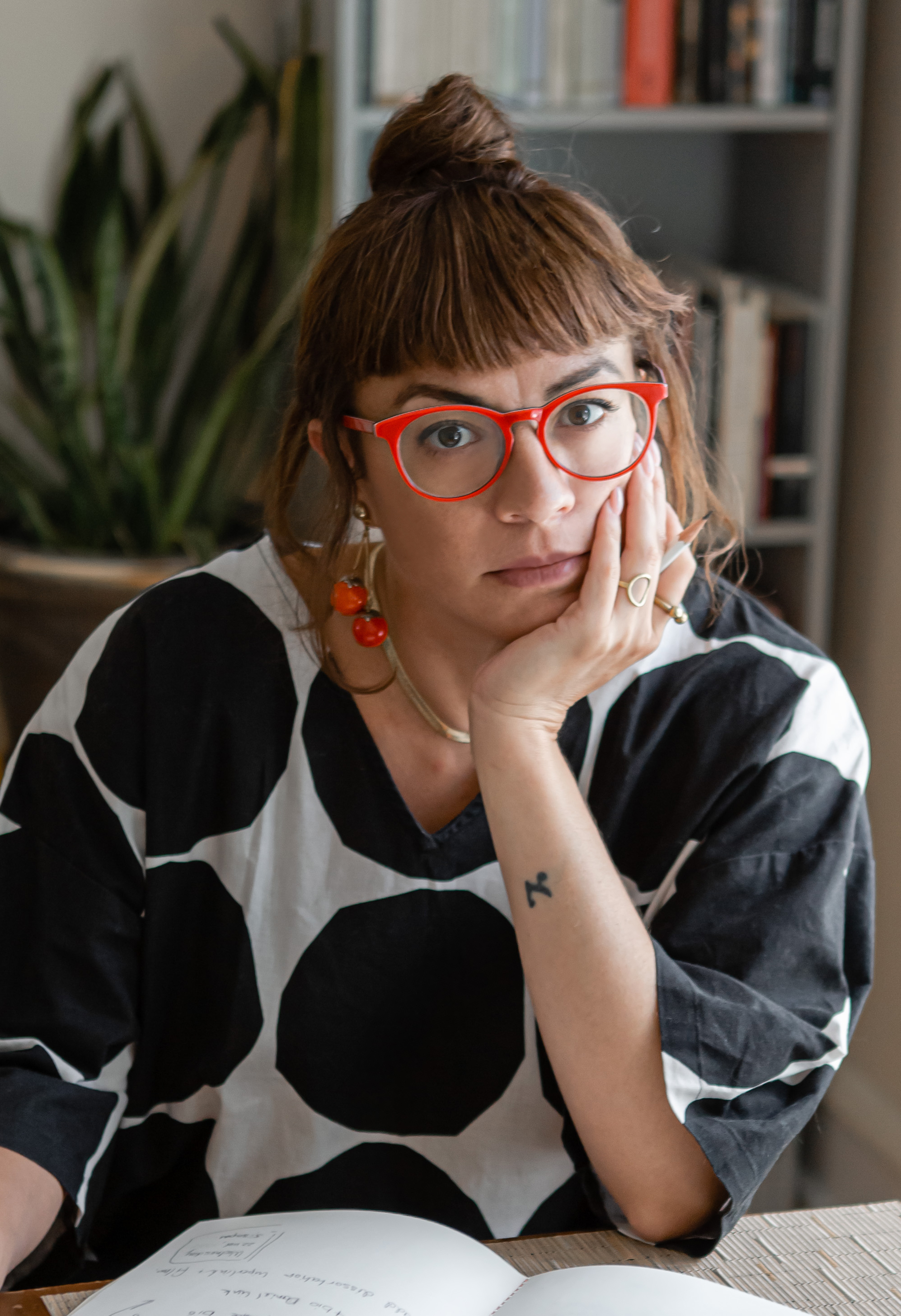 photographic portrait of white woman with red glasses and brown hair in a top bun, a bookshelf behind her