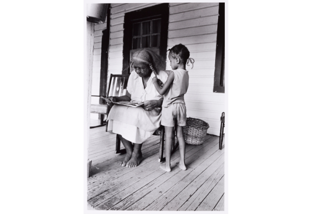 Older dark-skinned woman reading in a rocking chair on a porch while a child braids her hair