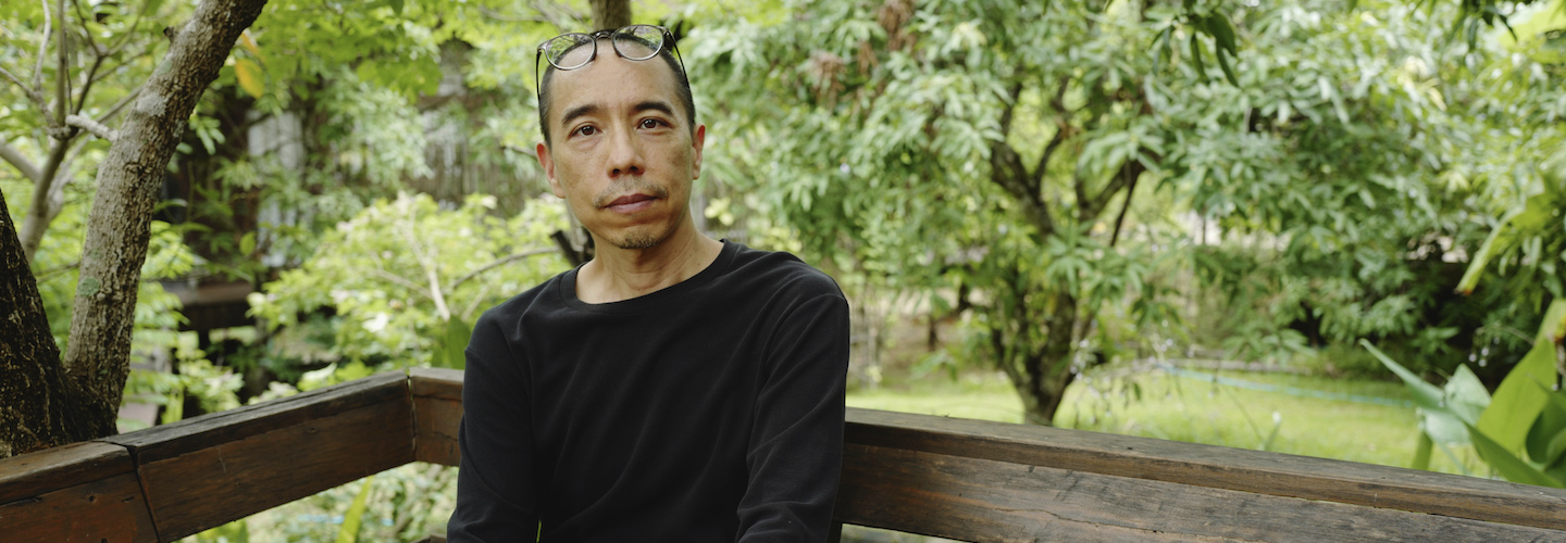 A portrait of Apichatpong Weerasethakul sitting in front of a grove of trees.