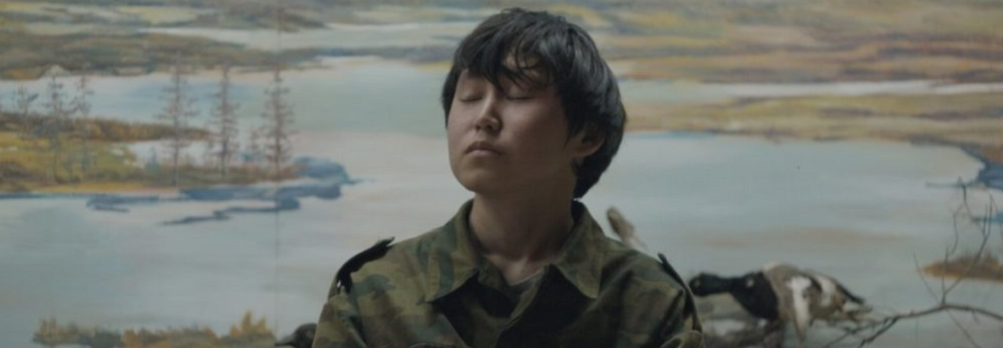 A young man with dark hair and medium skin tone sits meditatively, wearing a camouflage jacket, in front of a painted Siberian landscape. 