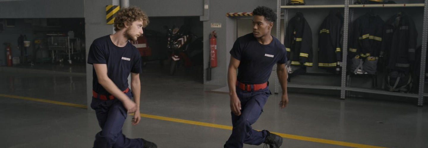 In a Fire Station, two young men in navy uniforms lock eyes and dance