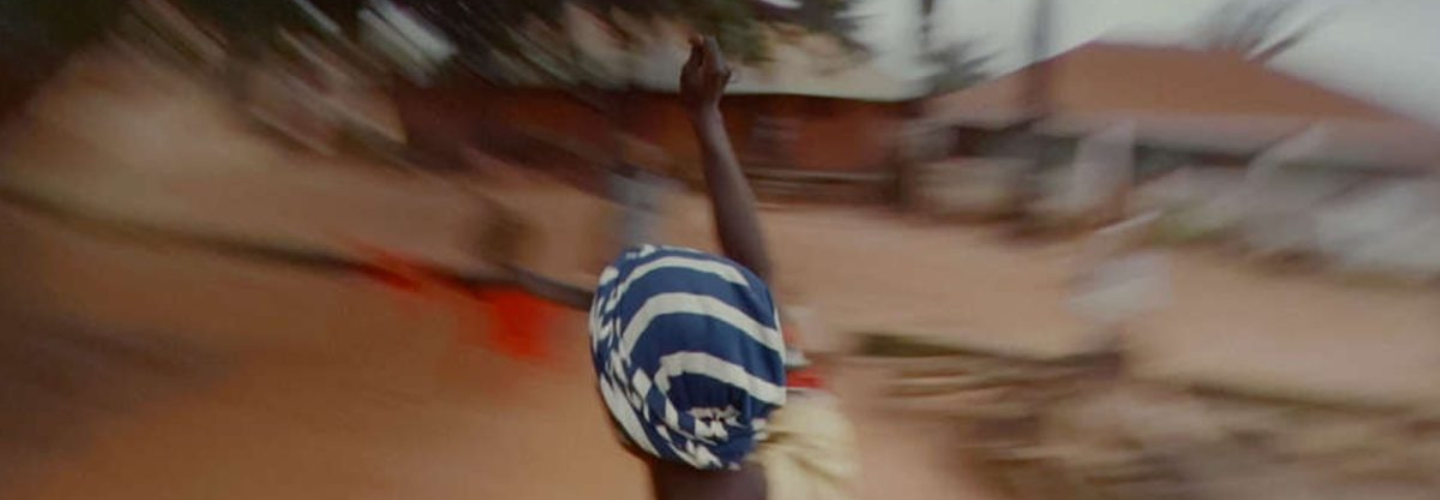 A person with a blue and white head wrap raises their arm surrounded by a blurry landscape 