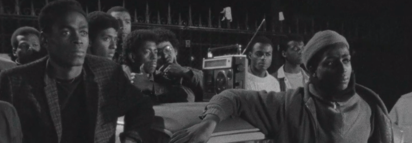A group of African-American men lean against a car, on the roof sits a boom-box.