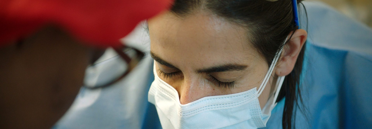 A woman with light skin and dark hair wearing a surgical mask and blue scrubs leans in toward another person with dark skin tone, glasses, and a red cap 