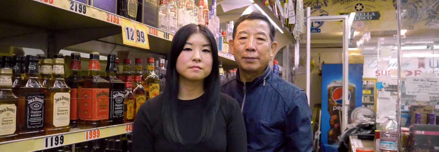 A daughter and father pose facing the camera with a backdrop of liquor store shelves