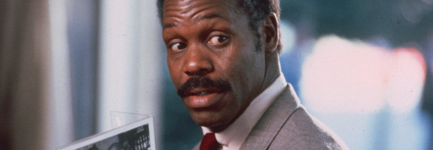 Danny Glover, wearing a grey suit, holds a photo and looks over his shoulder