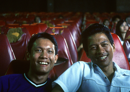 Two young men with medium skin tone sit side to side smiling in a movie theater