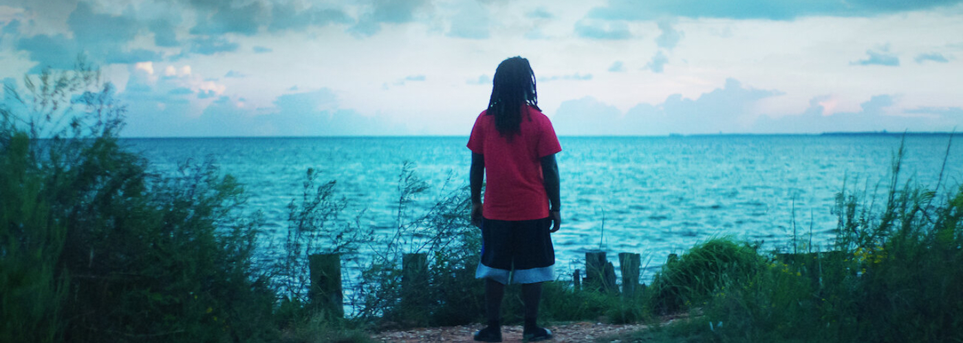 A man with dreadlocks in a red t-shirt and shorts stands looking out at the ocean.