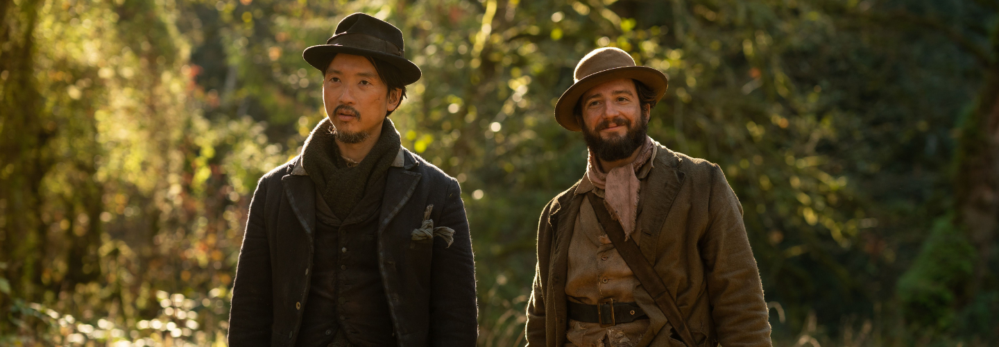 Two men dressed in frontiersmen clothing stand side by side in a vegetated setting