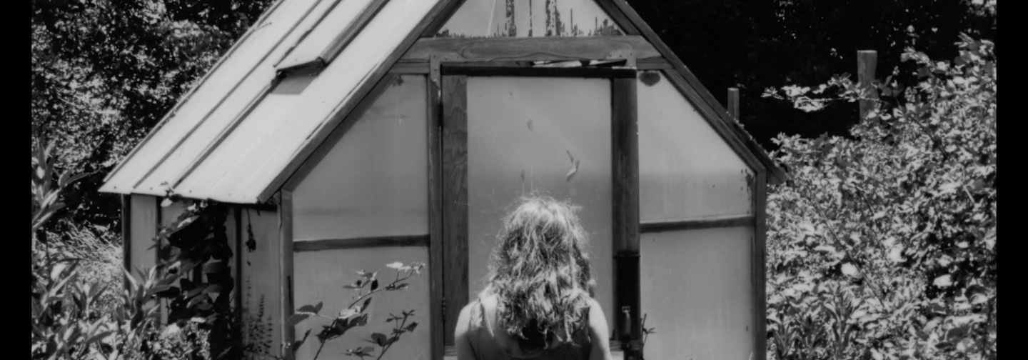 A long-haired figure standing in front of a small greenhouse in a verdant forest.