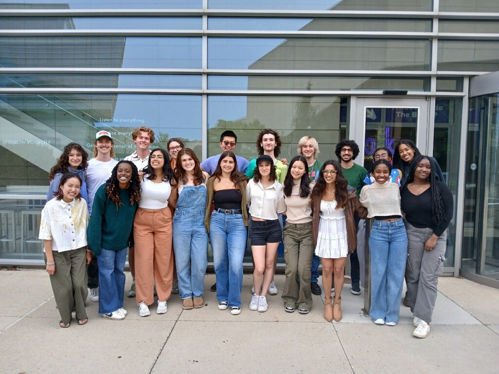 2 rows of people of diverse ethnicities and gender expressions, outside a glass building