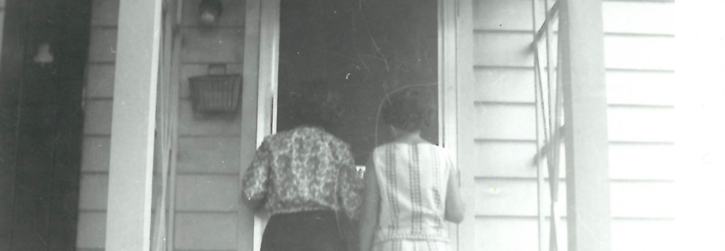 black and white photo of 2 women standing outside a house door, backs turned towards the camera