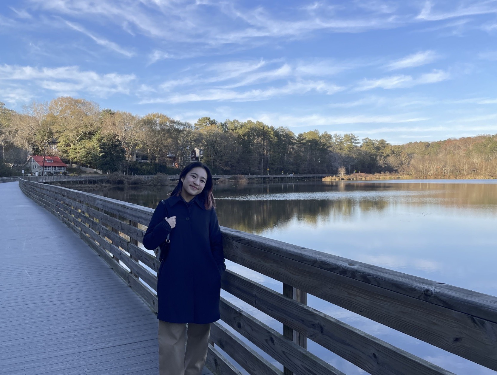 East Asian woman with navy coat, standing on a tree-lined river boardwalk