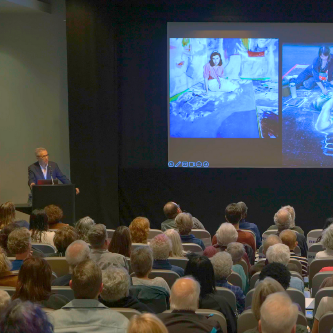 man stands at podium next to projector screen and is speaking to auditorium of listeners