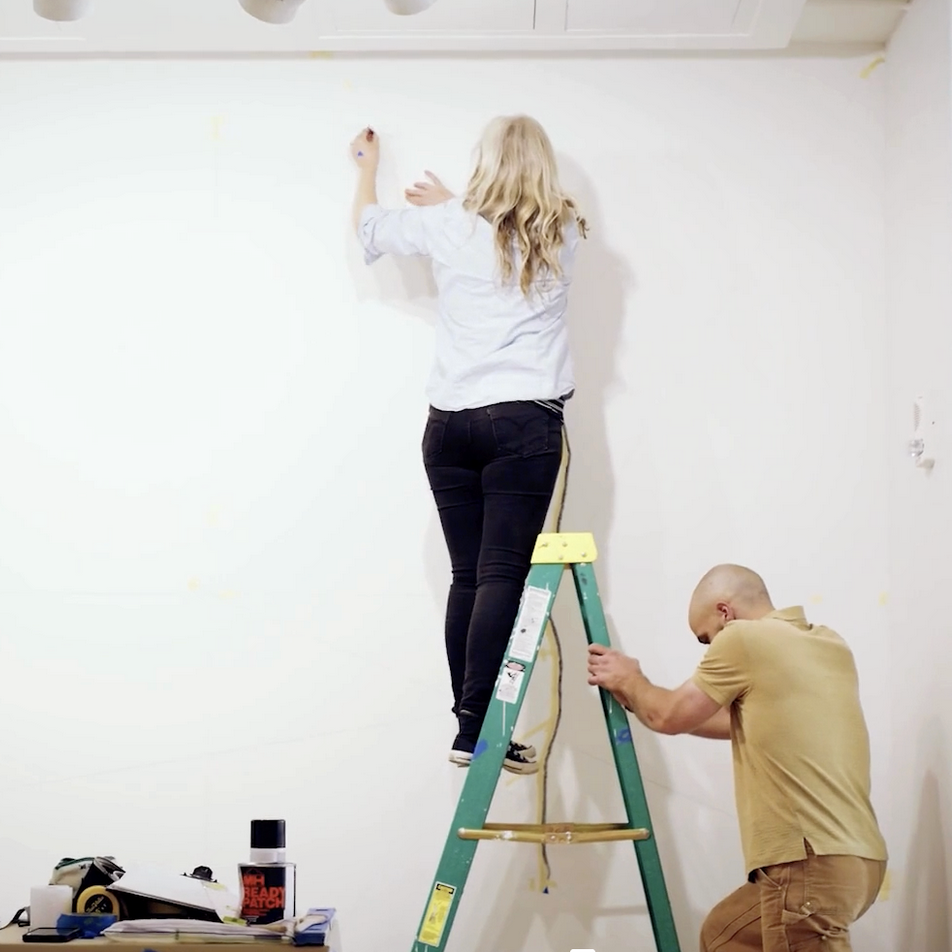 Woman stands on ladder, making marks on museum wall while man holds ladder still