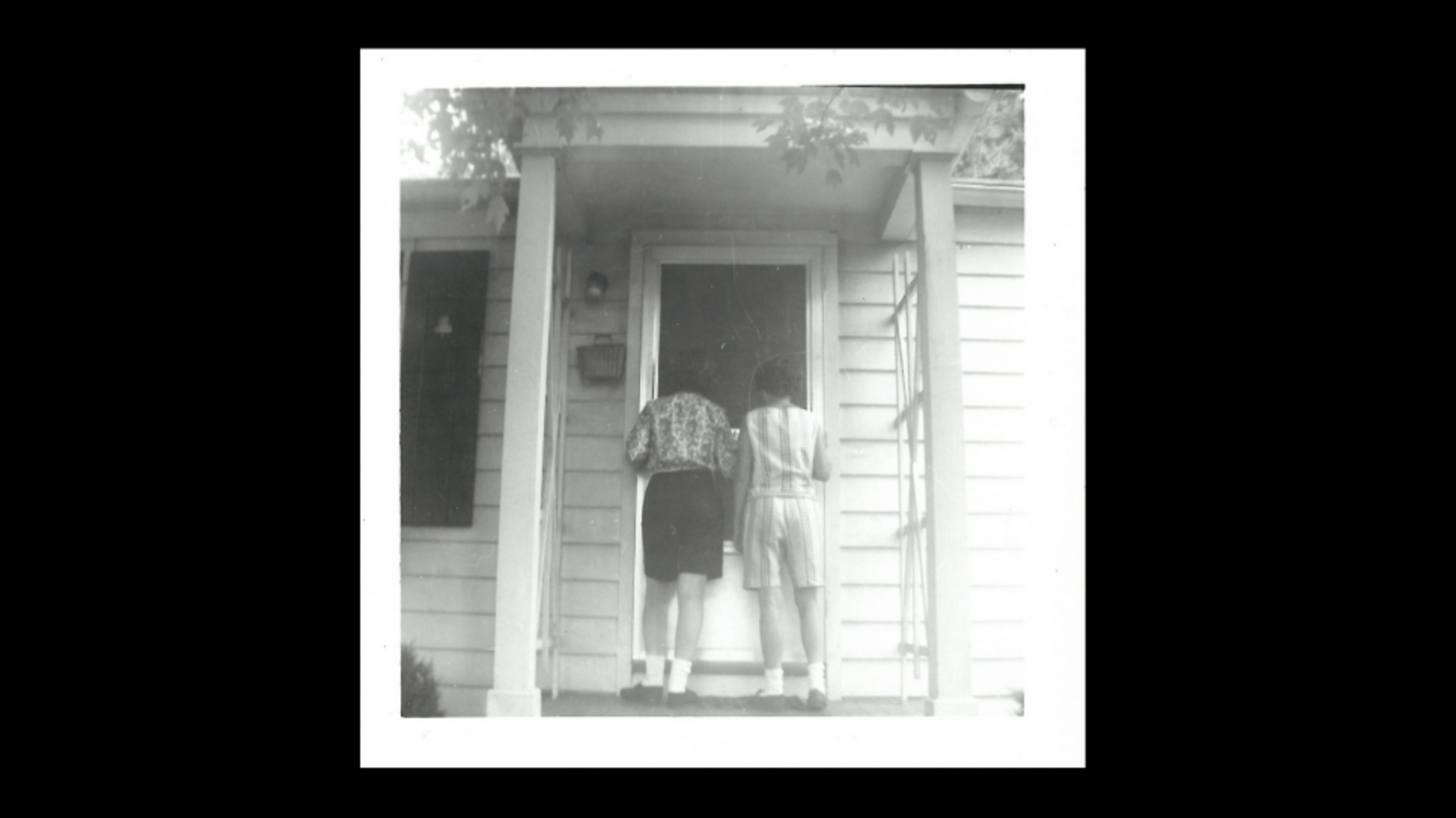square black-and-white photo on black background, of two people standing on a porch facing away from camera