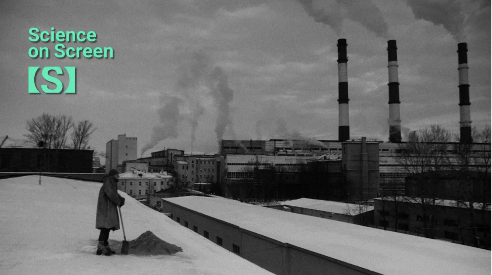 Black and white image of man with shovel standing on rooftop, with large smokestacks in background against cloudy sky